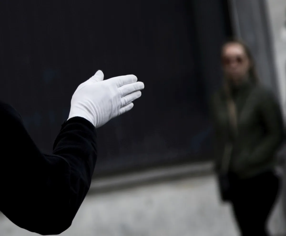 White-gloved hand extended toward a blurred woman against a dark wall